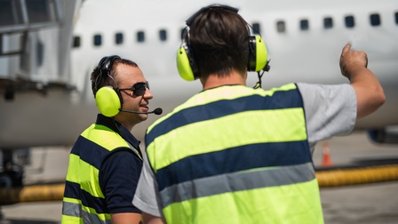 Zwei Personen in gelben Warnwesten mit gelben Kopfhörern in Seiten- und Rückenansicht, eine Person deutet auf Flugzeug, das im Hintergrund verschwommen zu sehen ist