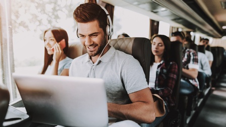Lächelnde Person mit Kopfhörern sitzt in Bus und blickt auf Laptop, ringsum weitere Personen sitzend