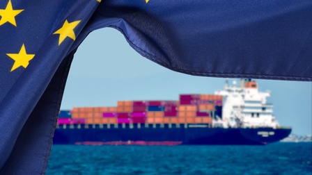 Im Vordergrund ist die Flagge der Europäischen Union, wie ein Vorhand zur Seite geschoben. Dahinter ist ein Containerschiff beladen mit zahlreichen Containern auf dem Meer, um Güter zu transportieren