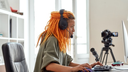 Person mit langen orangen Dreadlocks in Seitenansicht blickt auf Computermonitor, hat Kopfhörer aufgesetzt und bedient Maus und Tastatur, im Hintergrund weitere Mikrofon und Filmkamera