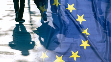 Im Hintergrund ist die Rückenansicht der Beine von vier Personen, die auf einer Straße gehen. Im Vordergrund ist die Flagge der europäischen Union darüber gelegt