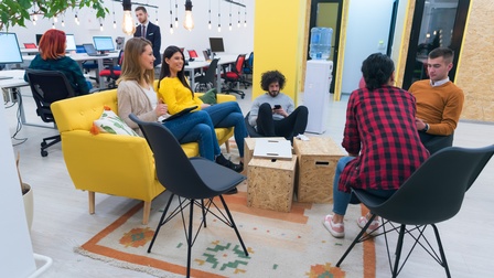 Personen sitzen in einem offenen, modernen und farbenfrohen Arbeitsraum und unterhalten sich, im Hintergrund stehen Schreibtische mit Computer