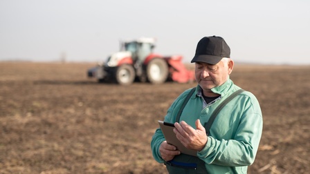 Person mit Kappe steht auf braunem Acker und blickt auf Tablet, im Hintergrund verschwommen Traktor