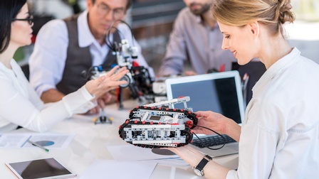 Personen sitzen an einem Tisch und betrachten Robotermaschinen, daneben stehen ein Laptop, ein Tablet sowie weitere Unterlagen