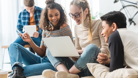 Drei lachende junge Personen auf Couch sitzend und auf Laptop blickend, im Hintergrund weitere Person auf Tisch sitzend