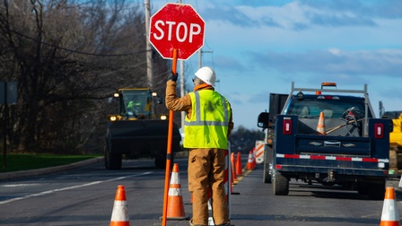 Eine Person mit einer Warnweste und Helm steht auf einer Straße und hält ein Stoppschild, im Hintergrund abgestellte Autos und ein fahrender Bagger.