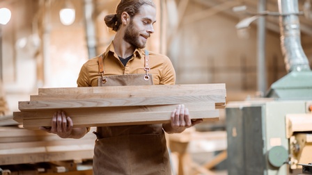 Lächelnde Person mit Bart in brauner Schürze blickt zur Seite und hält aufeinandergestapelte Holzbretter in Händen, im Hintergrund verschwommen Werkhalle
