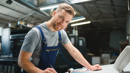 Bärtige sanft lächelnde Person in Arbeitslatzhose steht mit Stift an Tisch, Farbfächer vor sich liegend