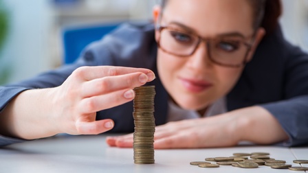 Person mit Brille lehnt an einem Tisch und formt Stapel mit Münzen, daneben liegen noch weitere Münzen wahllos am Tisch