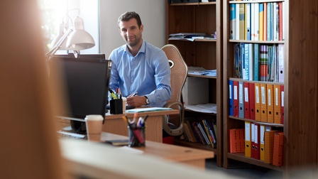 Lächelnder Unternehmer sitzt schreibend an Schreibtisch mit Bücherregal hinter sich