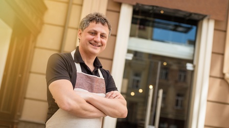 Lächelnde Person steht mit verschränkten Armen vor gläserner Eintrittstüre eines Geschäftslokals