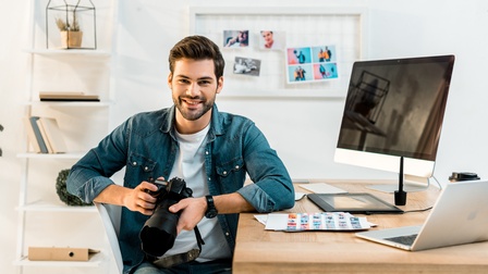 Eine Person mit einer Objektivkamera in den Händen sitzt seitlich an einem Tisch. Auf dem Tisch sind ein Bildschirm und ein Laptop. Vor der Person liegen Fotoabzüge