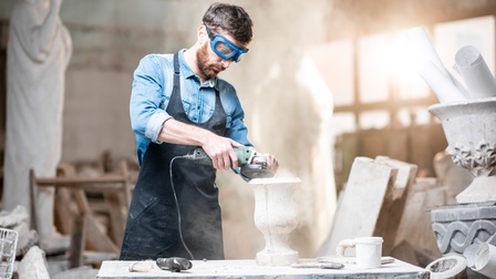 Bärtige Person mit Schutzbrillen in Schürze schleift mit elektrischer Handschleifmaschine Steingegenstand in Amphorenform auf Tisch aus Stein stehend, ringsum weitere Gegenstände und Skulpturen aus Stein