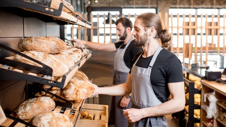 Zwei Personen hintereinander versetzt stehen vor Brotregalen und halten Brotlaibe in Händen