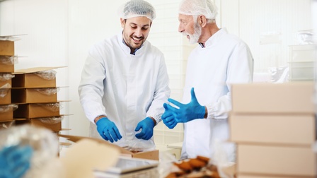 Personen mit Bart in weißer Arbeitskleidung, Haarnetzen und blauen Schutzhandschuhen verpacken Lebensmittel in einem Lager