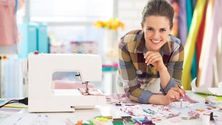 Lächelnde Person beugt sich über Tisch mit Stoffmustern, Schere, Köpfen und Zwirnen, neben ihr weiße Nähmaschine stehend, im Hintergrund verschwommen Stoffbahnen