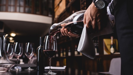 Zwei Hände einer Person halten einen Dekanter, mit dem sie eine dunkelrote Flüssigkeit in ein Weinglas einschenken, das auf einem Tisch steht. Dort steht eine weitere Weinflasche, leere Weingläser und ein leerer Dekanter