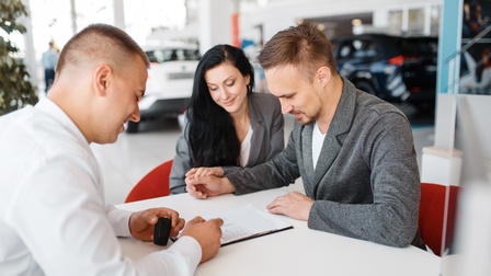 Paar blickt auf ein Dokument am Tisch in einer Empfangshalle, daneben sitzt eine weitere Person, die einen Autoschlüssel in Händen hält und auf das Dokument am Tisch zeigt, im Hintergrund stehen mehrere Autos