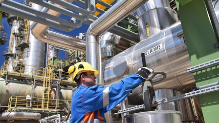 Person in blauem Overall mit gelbem Helm und Schutzbrillen dreht an Rad einer Gasanlage, ringsum verschiedene Leitungen und Container