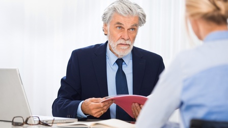 Eine Person hält die Bewerbungsmappe einer zum Bewerbungsgespräch eingeladenen Person, die gegenüber von ihr sitzt.
