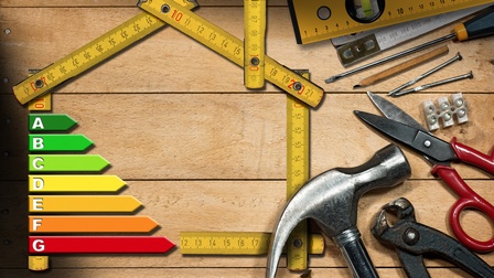 Topshot eines Maßbandes, das so drapiert ist, dass es die Umrisse eines Hauses ergibt. Links im Umriss ist ein Balkendiagramm zum Energiebedarf. Im unteren Bereich liegen verschiedene Werkzeuge wie Hammer, Bohrer, Nägel oder Zangen.