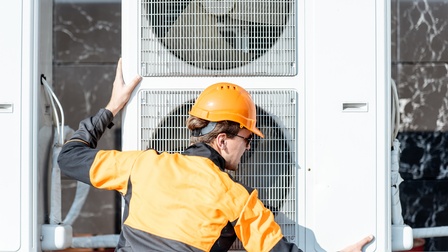 Rückenansicht einer Person mit schwarz-oranger Jacke, orangem Helm und Sonnenbrille, die neben einem Außengerät einer Klimaanlage steht und beide Hände auf dem Gerät hat