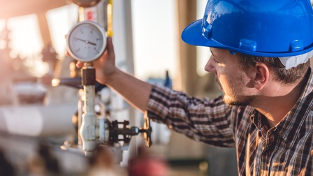 Person mit blauem Helm hält Hand an Manometer und blickt darauf.