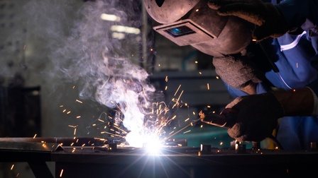 Eine Person mit Schweißhelm und Brille sowie dicken Arbeitshandschuhen beugt sich über eine Arbeitsplatte, um mit einem Schweißgerät etwas zu bearbeiten. Man sieht Funken und einen sehr hellen Punkt in der Bildmitte, wo geschweißt wird