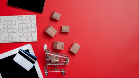 Auf einem roten Untergrund liegen in der linken Hälfte ein Smartphone, eine weiße Tastatur, ein Tablet, auf dem zwei Karten liegen, kleine Kartons und ein kleiner Einkaufswagen