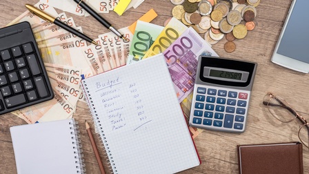 Text Budget mit Kalkulation auf einem Notizblock, ringsum liegen Euroscheine und Euromünzen sowie Taschenrechner, Stifte, ein Smartphone, Notizbücher und eine Brille auf einem Holzuntergrund