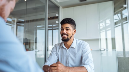 Eine Person spricht an einem Tisch in einem Besprechungszimmer sitzend mit einer ihr gegenüber sitzenden Person