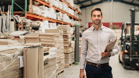 Porträt einer Person mit Klemmbrett in der rechten Bildhälfte. Links neben der Person sind viele, eher schmale übereinander gestapelt. Im Hintergrund ist in der linken Hälfte ein großes, hohes Regal,  in dem verschiedene Objekte sind und ein Gabelstapler.