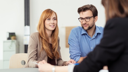 Zwei Personen sitzen an Tisch, eine Person lächelt sanft, die andere trägt Brillen, im Vordergrund verschwommen weitere Person