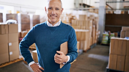 Person mit Pullover steht mit einem Klemmbrett und Kugelschreiber freudig in einer Lagerhalle