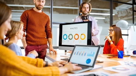 Personen an einem Tisch in einem modernen Büro unterhalten sich während Analysen und Grafiken auf einem Bildschirm sowie auf einem Laptop visualisiert sind