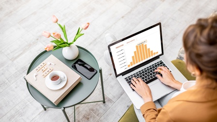 Person sitzt auf einem Sofa und blickt auf ihr Notebook mit einer Tabellen-Grafik auf ihrem Schoß, daneben steht ein Beistelltisch mit einer Blumenvase, einem Work at Home Buch sowie einem Smartphone