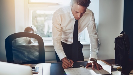 Person in Businesskleidung steht vor einem Schreibtisch und unterzeichnet ein Dokument, daneben steht eine Aktentasche sowie ein Glas Wasser 