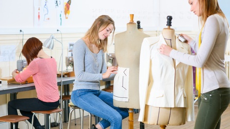 Zwei Personen stehen jeweils neben einer Modepuppe. Die rechte Person hat ein Maßband um den Hals. An einem Tisch in der linken Bildhälfte sitzt eine Person an einer Nähmaschine. An der Wand hängen Papiere mit Skizzen.