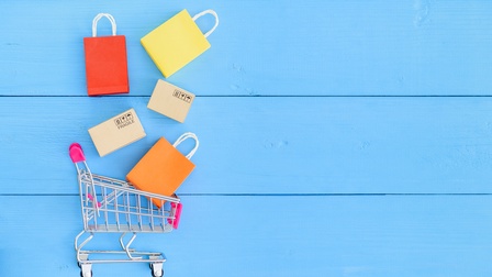 Auf einer blauen Unterfläche liegt in der linken Hälfte ein Miniatur-Einkaufswagen. Über dem Einkaufswagen liegen drei bunte Einkaufstaschen und zwei Kartons. Auf einem Karton steht fragile.