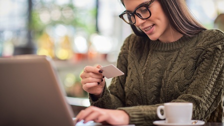 Person mit Brille und grünem Strickpullover sitzt mit einer Tasse vor einem Laptop und hält eine EC-Karte in der Hand während im Laptop etwas eingegeben wird