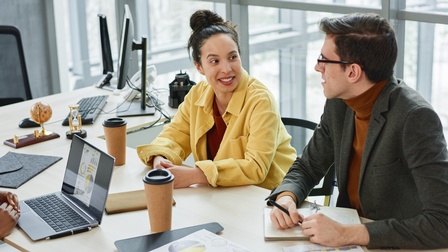 Zwei Personen an Arbeitstisch sitzend in Unterhaltung miteinander, am Tisch verschiedene Arbeitsutensilien, Dokumente mit statistischen Grafiken und Kaffeebecher