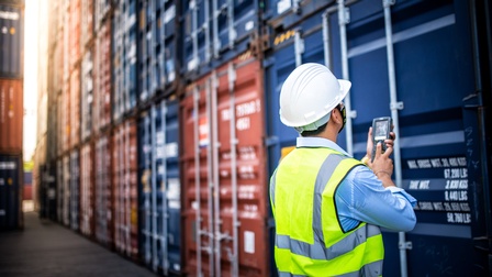 Rückenansicht einer Person mit Sicherheitsweste und weißem Helm, die mit den Händen einen Scanner hält. Direkt vor der Person steht ein blauer Container. Darüber und daneben sind weitere Container gestapelt.