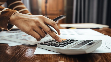 Eine Person tippt mit der rechten Hand auf einem Taschenrechner, der auf einem Tisch steht. Ihre linke Hand hält ein Smartphone. Auf dem Tisch sind verschiedene Zettel