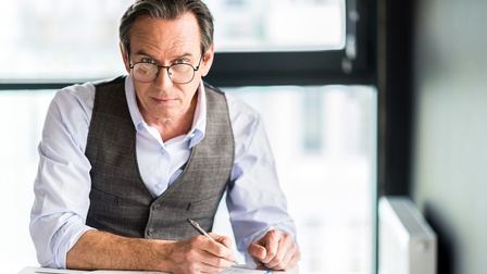 Person mit Brille mittleren Alters sitzt in Businesskleidung an einem Schreibtisch und bearbeitet ein Dokument mit einem Stift, im Hintergrund ist eine helle Fensterfront 