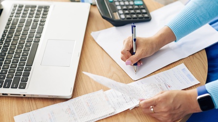 Topshot der Hände einer Person, die mit der linken Hand eine Rechnung hält. Unter dieser sind weitere Rechnungen. Rechts daneben notiert die Person mit einem Stift etwas auf einem Zettel. Daneben steht ein Taschenrechner