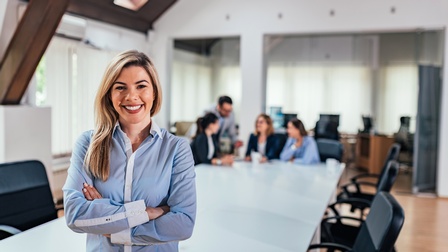 Porträt einer lächelnden Person in einem Hemd und mit verschränkten Armen, die vor einem sehr großen Tisch sitzt. Am anderen Ende des Tisches sitzen drei Personen. Eine weitere steht daneben. Die vier Personen reden miteinander