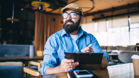 Person mit Brillen, Bart  und Cap in Lokal sitzend macht Notizen auf Clipboard