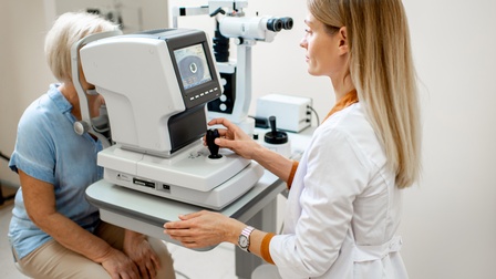 Person in weißer Arbeitskleidung bedient ein digitales Mikroskop und betrachtet die Augen einer älteren Person mit weißen Haaren