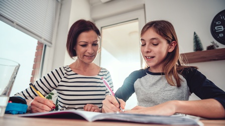 Frontalaufnahme einer erwachsenen Person und eines Kindes, die nah nebeneinander hinter einem Tisch sitzen. Vor dem Kind ist ein aufgeklapptes Heft, auf das beide blicken. Das Kind hält einen Stift darauf