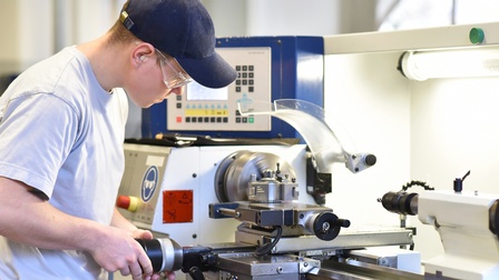 Eine Person steht vor einer CNC-Drehmaschine und bedient diese konzentriert. Sie trägt eine Schirmkappe.